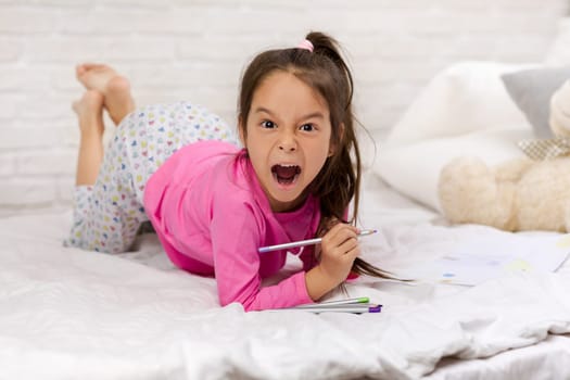 cute little girl drawing pictures while lying on bed. Kid painting at home