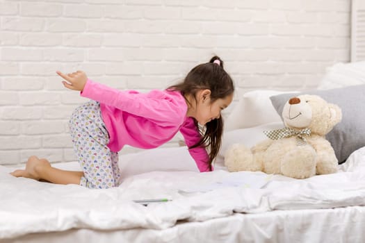 adorable little multinational child girl playing with teddy bear in bed in morning