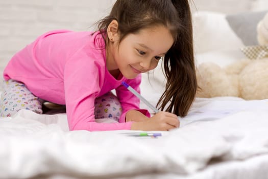 funny little girl drawing pictures while lying on bed. Kid painting at home