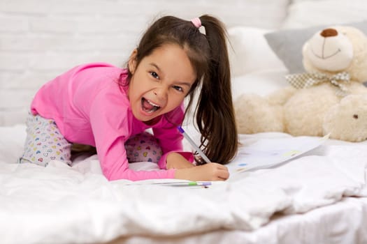 funny little girl drawing pictures while lying on bed. Kid painting at home