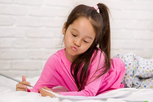 cute little girl drawing pictures while lying on bed. Kid painting at home