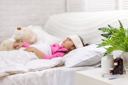 Sick little child girl lying in bed. Cold flu season. focus on medication and pill bottle