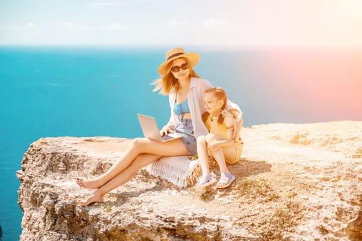 Freelance woman with her daughter working on a laptop by the sea, typing on the keyboard, enjoying the beautiful view, highlighting the idea of remote work