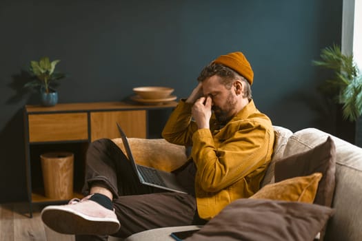 Sad and tired man is depicted sitting on a sofa at home. Overwhelmed by bad news or challenges, he holds his head with closed eyes in his hand, conveying a sense of emotional distress. High quality photo