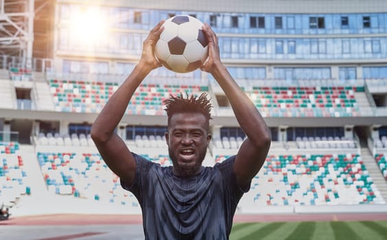 Young confident african american guy holds soccer ball in hands outside, on stadium and looks up.