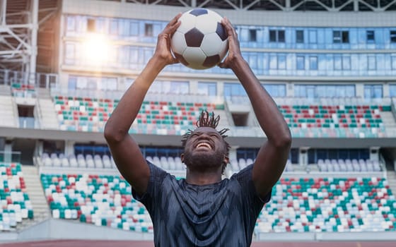 Young confident african american guy holds soccer ball in hands outside, on stadium and looks up.