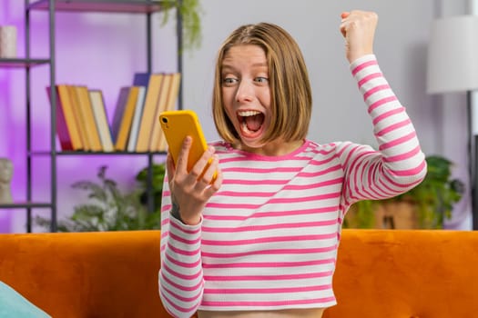 Happy woman use mobile smartphone typing browsing say Wow yes found out great big win good news celebrate lottery jackpot doing winner gesture. Portrait of brunette girl at home in room sits on sofa