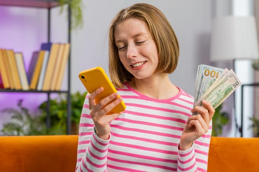 Planning family budget. Smiling young brunette woman counting money cash, use smartphone calculate domestic bills at home room. Girl satisfied of income and saves money for planned vacation, gifts