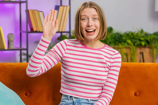 Hello. Brunette woman smiling friendly at camera, waving hands gesturing hello, hi, greeting or goodbye, welcoming with hospitable expression at home. Portrait of girl sitting on sofa in living room