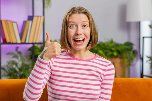 Like. Happy excited brunette woman looking approvingly at camera showing thumbs up, like sign positive something, good great news, positive feedback. Young girl sitting on couch at home living room