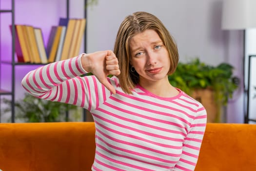 Dislike. Portrait of upset brunette woman showing thumbs down sign gesture, expressing discontent, disapproval dissatisfied bad work at modern home apartment indoors. Displeased girl in room on sofa