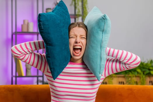 Repair work at neighbours. Irritated woman girl relaxing on couch cover ears with pillows annoyed by noisy neighbors suffer from headache wish silence. Thin walls at home flat without sound insulation