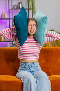 Repair work at neighbours. Irritated woman girl relaxing on couch cover ears with pillows annoyed by noisy neighbors suffer from headache wish silence. Thin walls at home flat without sound insulation