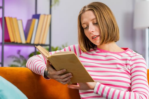 Young brunette woman relaxing interesting book turning pages smiling enjoying literature taking a rest on comfortable sofa. Peaceful smiling cheerful girl at home apartment living room sits on couch
