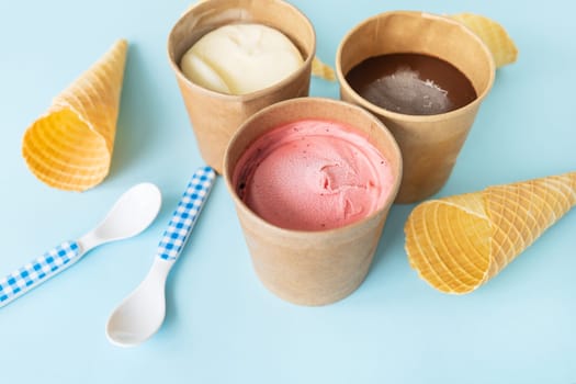 Ice cream in a craft box with different flavors - strawberry, chocolate, vanilla stands on a blue background along with a waffle cone and a checkered spoon. View from above