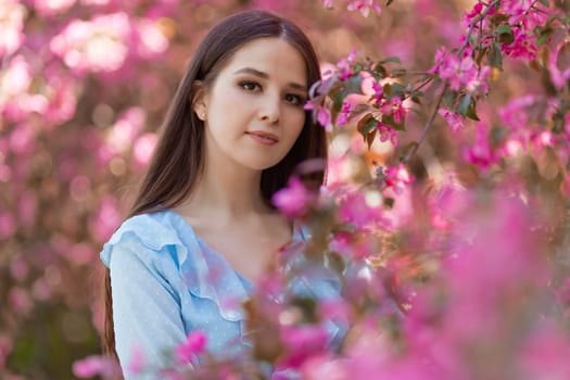 Portrait of adorable brunette girl with long hair is standing near a pink blooming apple tree, in the summer in the park. Close up