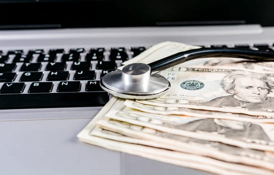 Stethoscope on top of money on laptop keyboard. Stethoscope on dollar bills on top of laptop keyboard