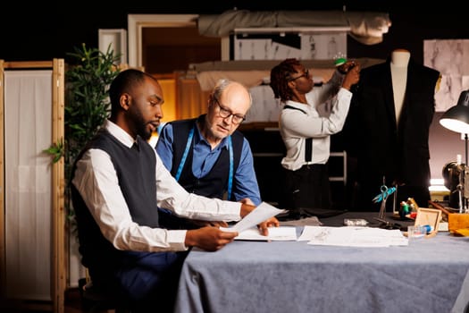 Stylish customer in tailoring shop asking for suit customizations, discussing next steps with expert suitmaker over fashion sketches. Client looking at clothing designs, choosing desired alterations