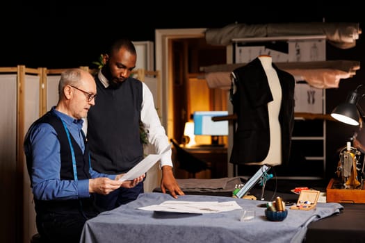 Skilled tailors comparing different fashion sketches attire combinations in atelier workspace. Dressmaker and apprentice working on clothing alterations, preparing for upcoming collection release