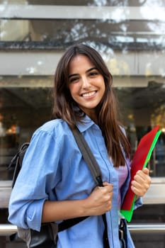 Vertical portrait of young female caucasian college student looking at camera. Young woman with backpack standing outside university building holding folders. Education concept.