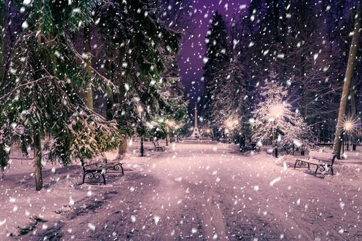 Snowfall in a winter park at night with christmas decorations, lights and  pavement covered with snow. Falling snow.