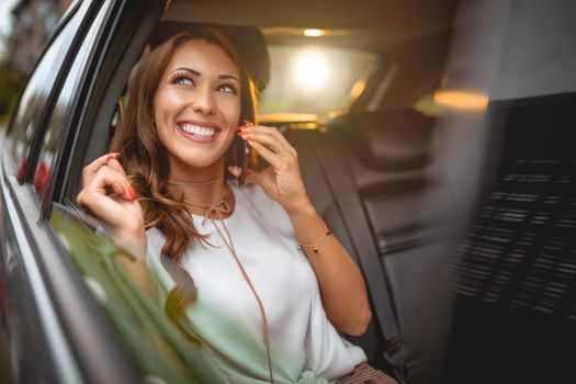 Young smiling woman holding smartphone in her hand and talking on the back seat in car.