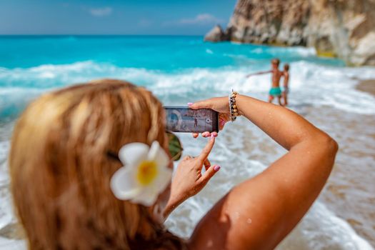 Woman with smartphone takes a photo of her loving sons in sea waves on the tropical beach. Selective focus. Focus on photo on the phone.