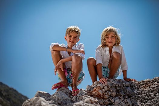 Two cute boys are sitting on the sea rock on the beach and looking down at camera.