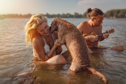 A young happy woman is sitting in the water on the river beach and petting their dog. In the background there is a musician sitting in the water and  playing on ukulele.  