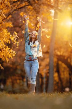Cheerful young beautiful woman going alone in the park and throws leaves up in golden sunny autumn.