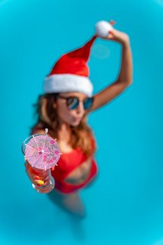 Young beautiful woman near the swimming pool in Santa Claus hat celebrating New Year and Christmas in hot country with glass of cocktail.
