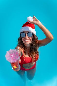 Young beautiful woman in the swimming pool in Santa Claus hat celebrating New Year and Christmas in hot country with glass of cocktail.