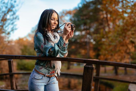 Woman photographer holding professional digital camera and taking photo on the bridge in autumn city park.  