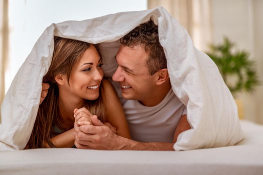 Beautiful young loving couple looking each other with love and having lazy morning in bed covered with blanket.