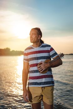 Happy young musician has a great time at the beach. He is standing by the water, holding ukulele and singing. Sunset over water.