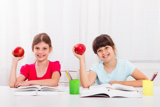Two happy schoolgirls with apple, books and pencils at table