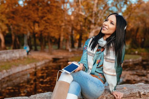 Young woman listening music on a smartphone with headphones on her ears and sitting in a park in a wonderful autumn day.