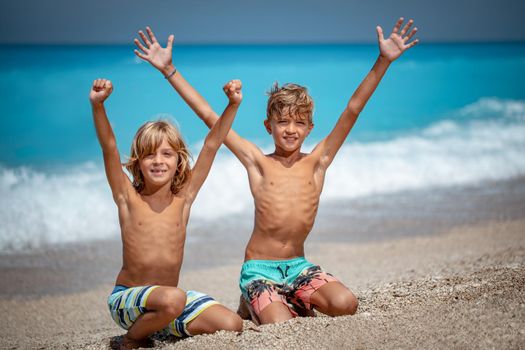 Two cute brothers are sitting on the beach and smiling looking at camera. Their arms are wide open.  
