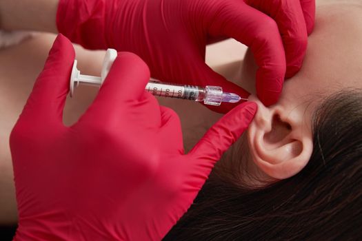 cosmetologist makes the injections procedure for smoothing wrinkles and against flabbiness of the skin on earlobe of a beautiful, young woman