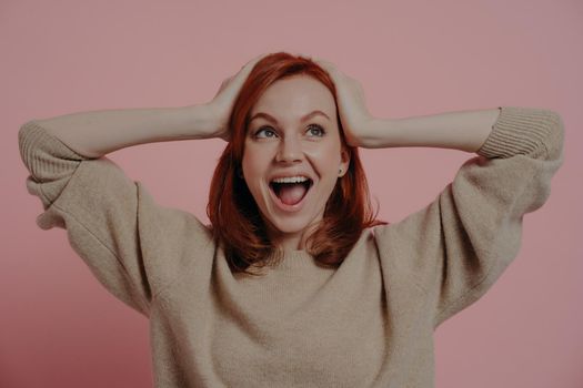 Surprised shocked european female holding head in hands and looking upside with excited expression and opened mouth, hearing about big sale prices, standing in studio isolated over pink background