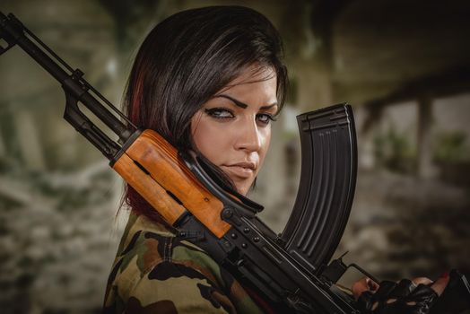 Attractive dark-haired girl in camouflage shirt hold machine gun leaning on the shoulder and with a dangerous expression on her face looking at the camera.