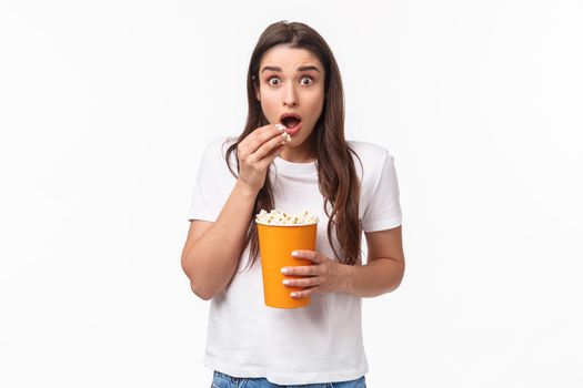 Entertainment, fun and holidays concept. Portrait of astonished and excited impressed young girl watching awesome movie, look astonished eating popcorn, pop eyes at tv screen, white background.