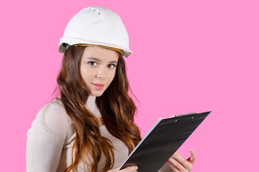 A young female engineer in a helmet holds her project in her hands on the pink background.