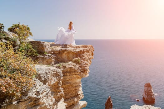 Woman in a white dress on the sea. Side view Young beautiful sensual woman in white long dress posing on a rock high above the sea at sunset. Girl in nature against the blue sky.