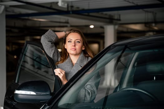 Happy woman car. she stands next to the car in the underground parking. Dressed in a gray coat, holding a glass of coffee in her hands, a black car
