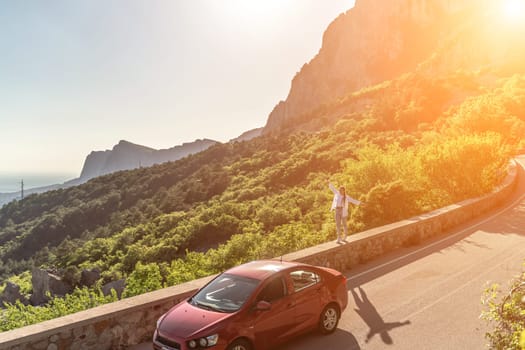 Mountain road woman. She walks along a parapet road in the middle of a mountainous area, a car is parked nearby. She is dressed in jeans and a white shirt, her hair is braided