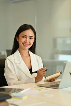 Portrait of confident asian woman manager sitting at office desk with laptop and paperworks.