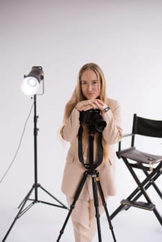 A woman videographer, a blonde smiling with a camera Lumix GH 5 on a tripod in the studio. Wearing a formal nude pantsuit on a white isolated background, vertical