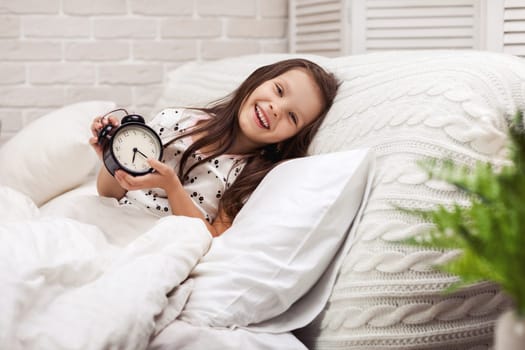 cute happy little child girl in pyjamas with clock in bed. good morning