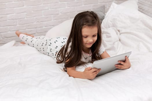 cute little child girl lies in bed uses digital tablet. child playing on tablet pc.